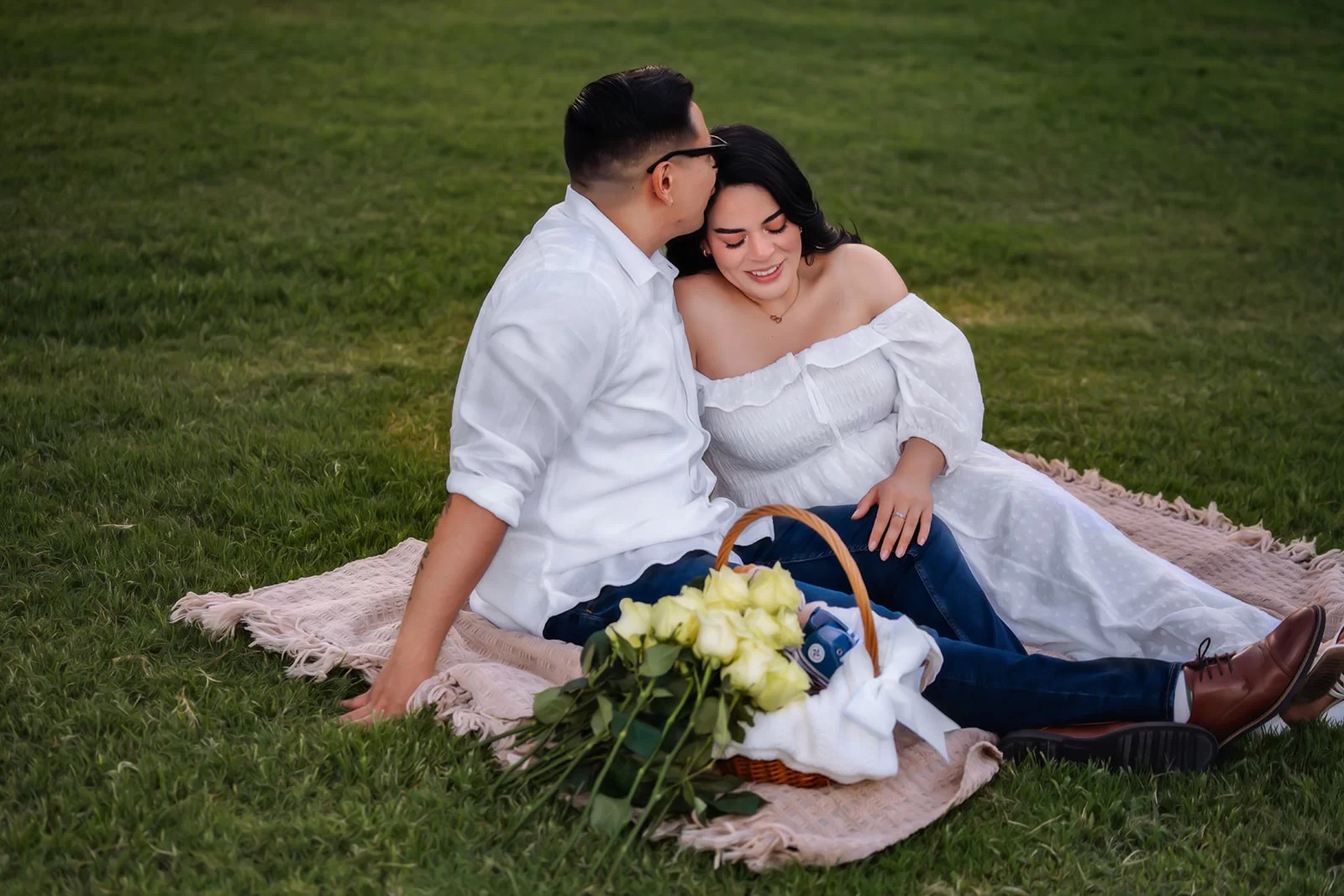 Couple sits in the grass kissing and smiling for their maternity photos.